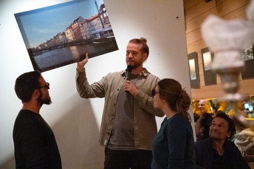 Simon Kimmel steht auf einem Speaker Podest, in der einen Hand ein Mikro in der Anderen ein Bild von Copenhagen. Er versucht dem Publikum beispielhaft Virtual Reality zu erklären